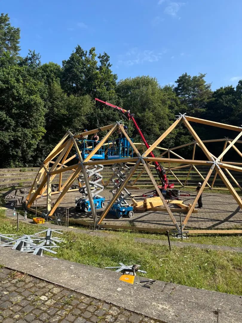 The half assembled wooden framework of the dome in the Theatron in the Ostpark, with two lifts, a crane and workers on the platform