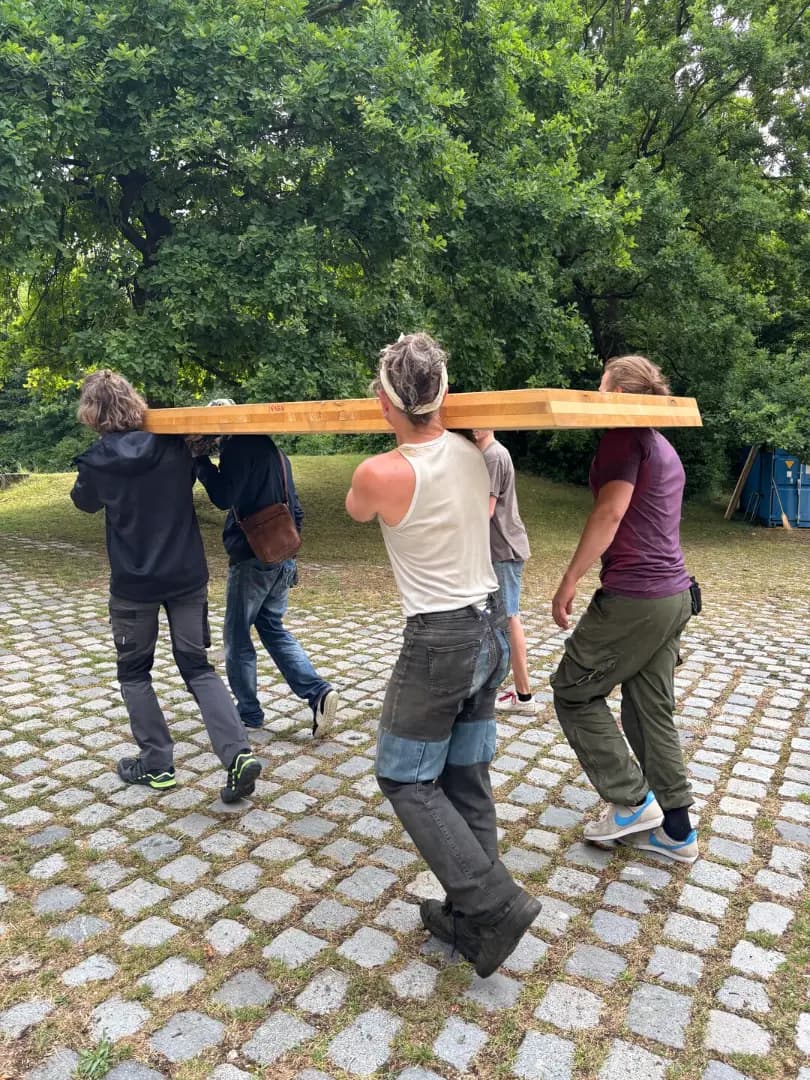 Four people carrying a long wooden beam together across the paving to the build site