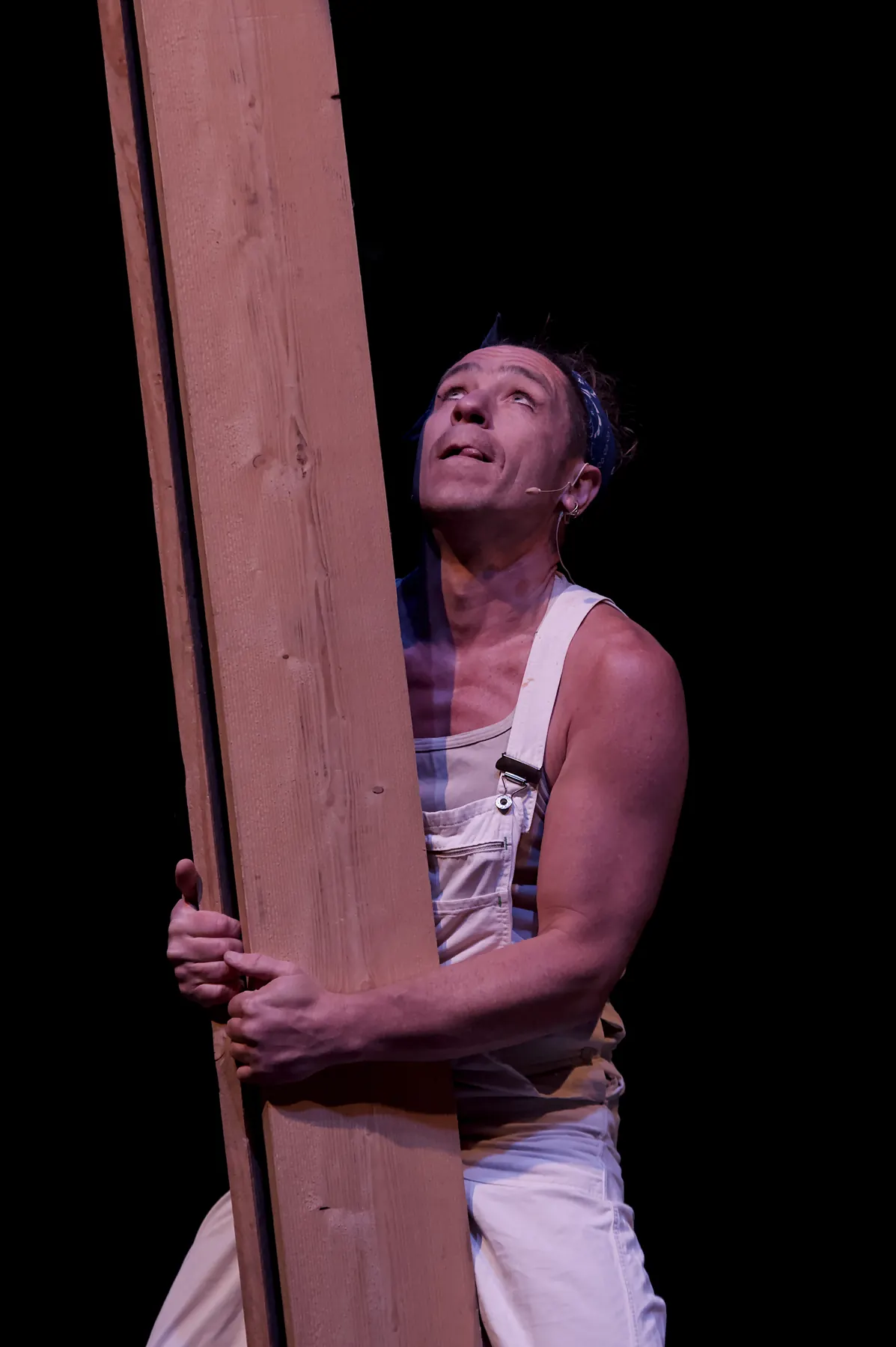 Artist in white dungarees balancing an upright wooden plank and looking up along it
