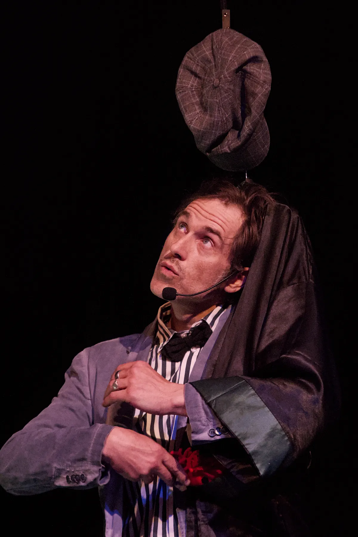 A clown in a grey jacket looking up at a flat cap hanging in the air above him
