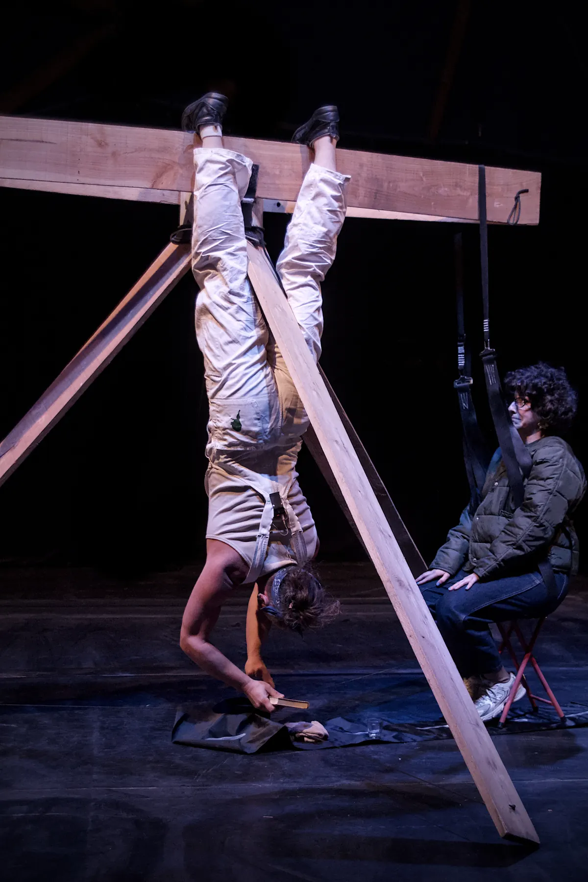 Artist upside down on a tilted wooden plank, a person sitting on a folding chair beside him