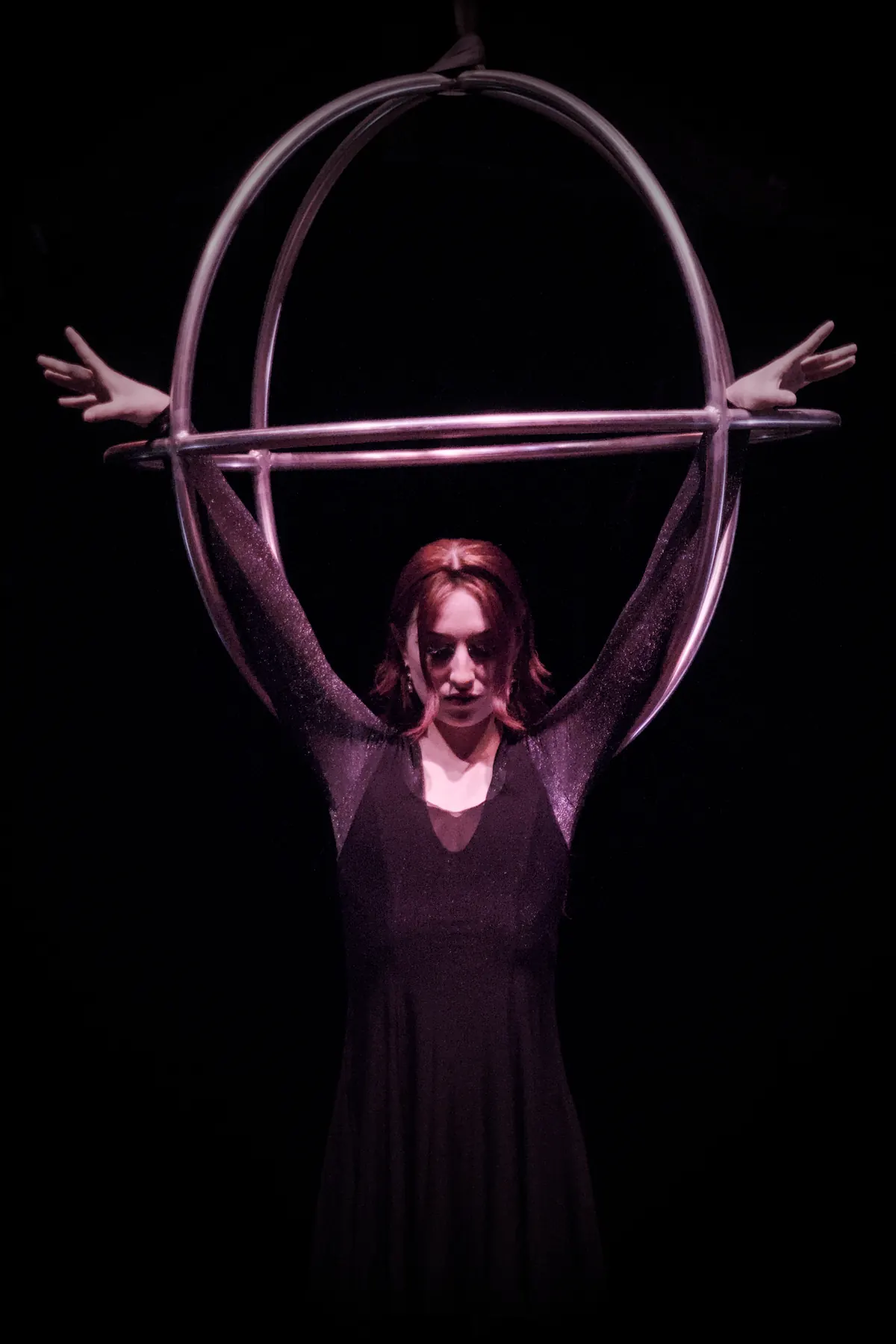 Woman in a black dress standing inside an apparatus of two crossed hoops, arms stretched out sideways