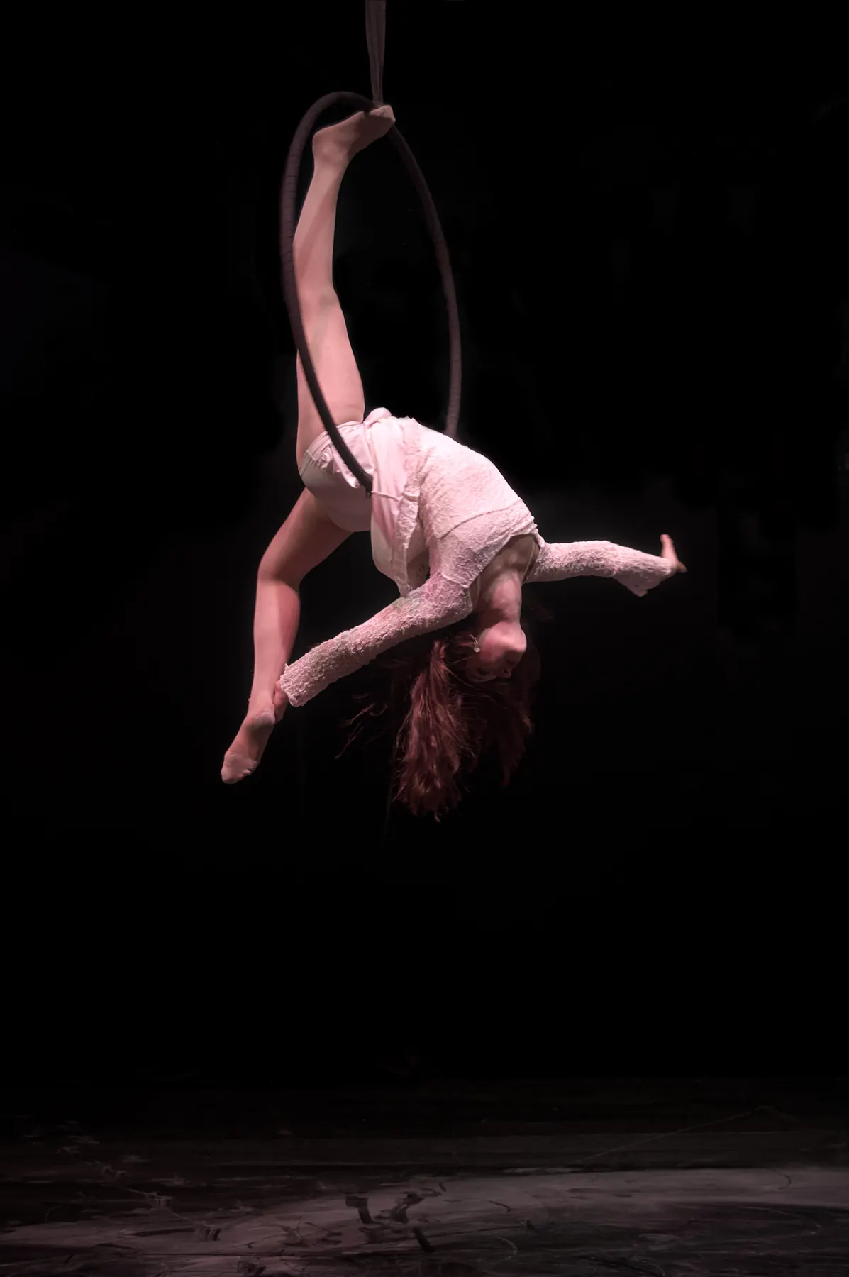 Artist in a pale dress hanging upside down from an aerial hoop above the dark stage