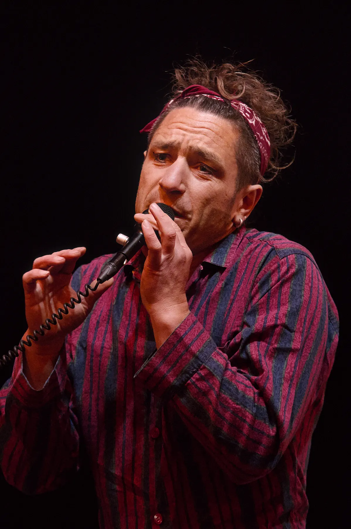 Musician in a red and blue striped shirt singing close into a handheld microphone