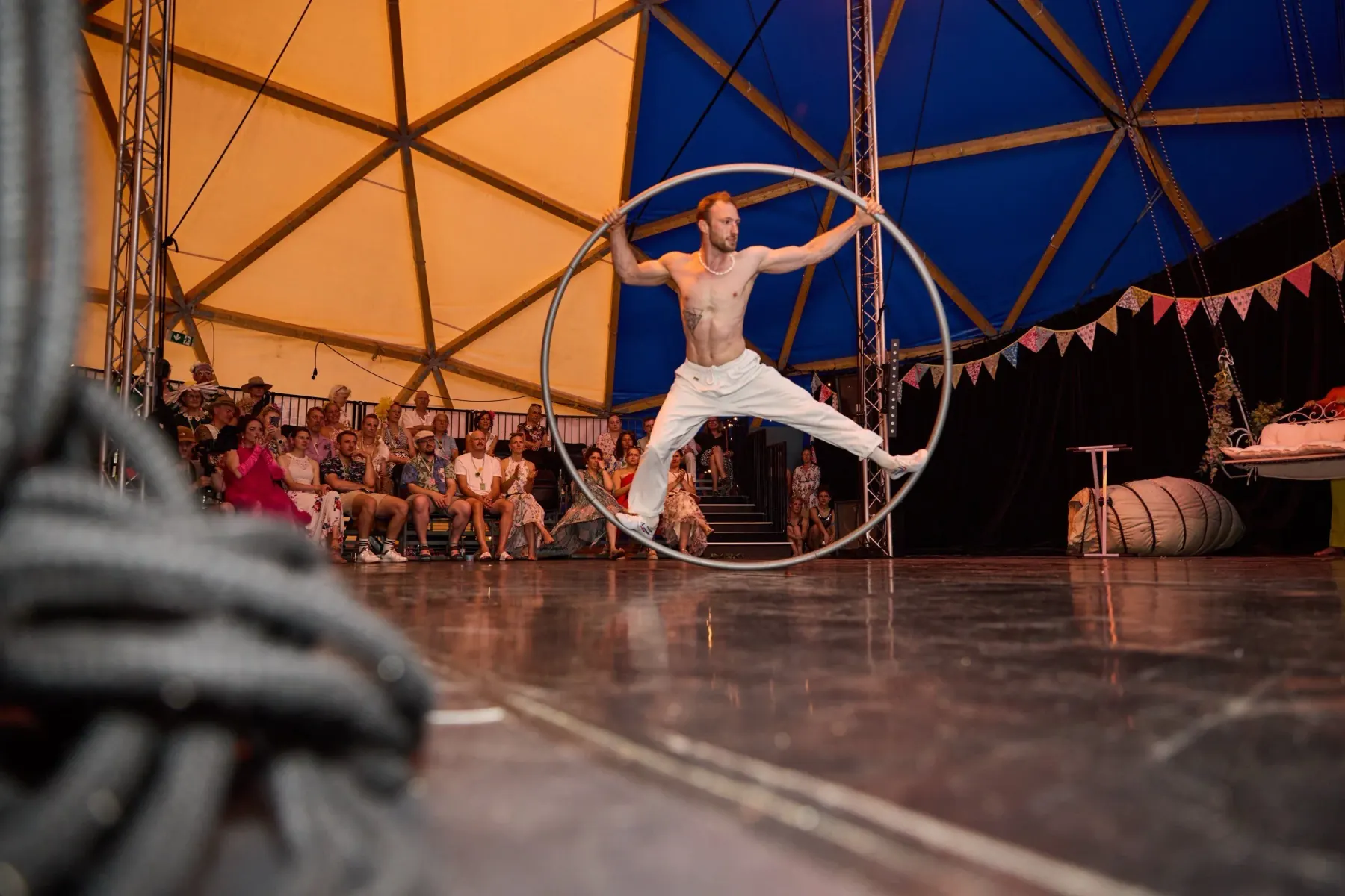 Artist standing wide-legged inside a Cyr wheel on stage, the blue canopy of the dome and the audience behind