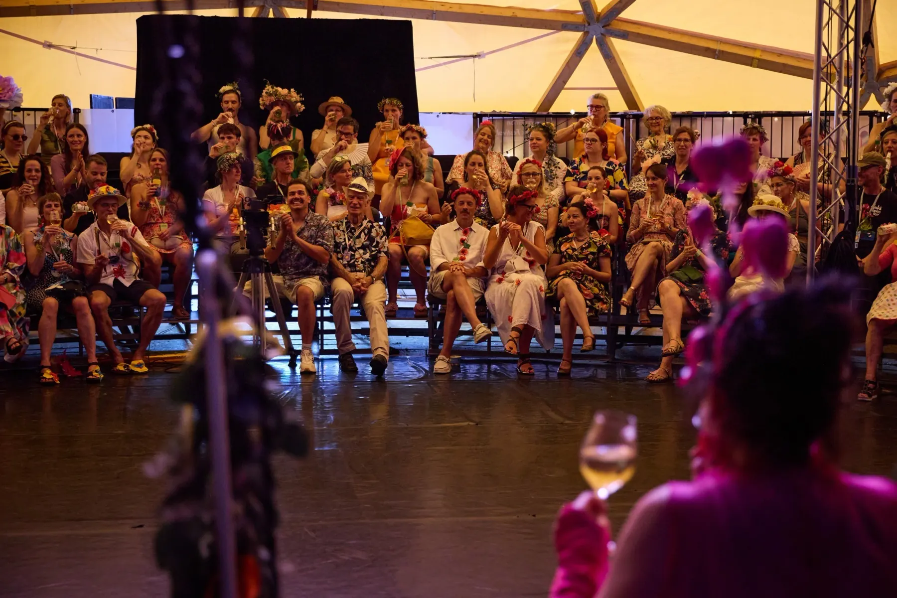 Full rows of spectators inside the dome on a summer day, many guests wearing flowers in their hair