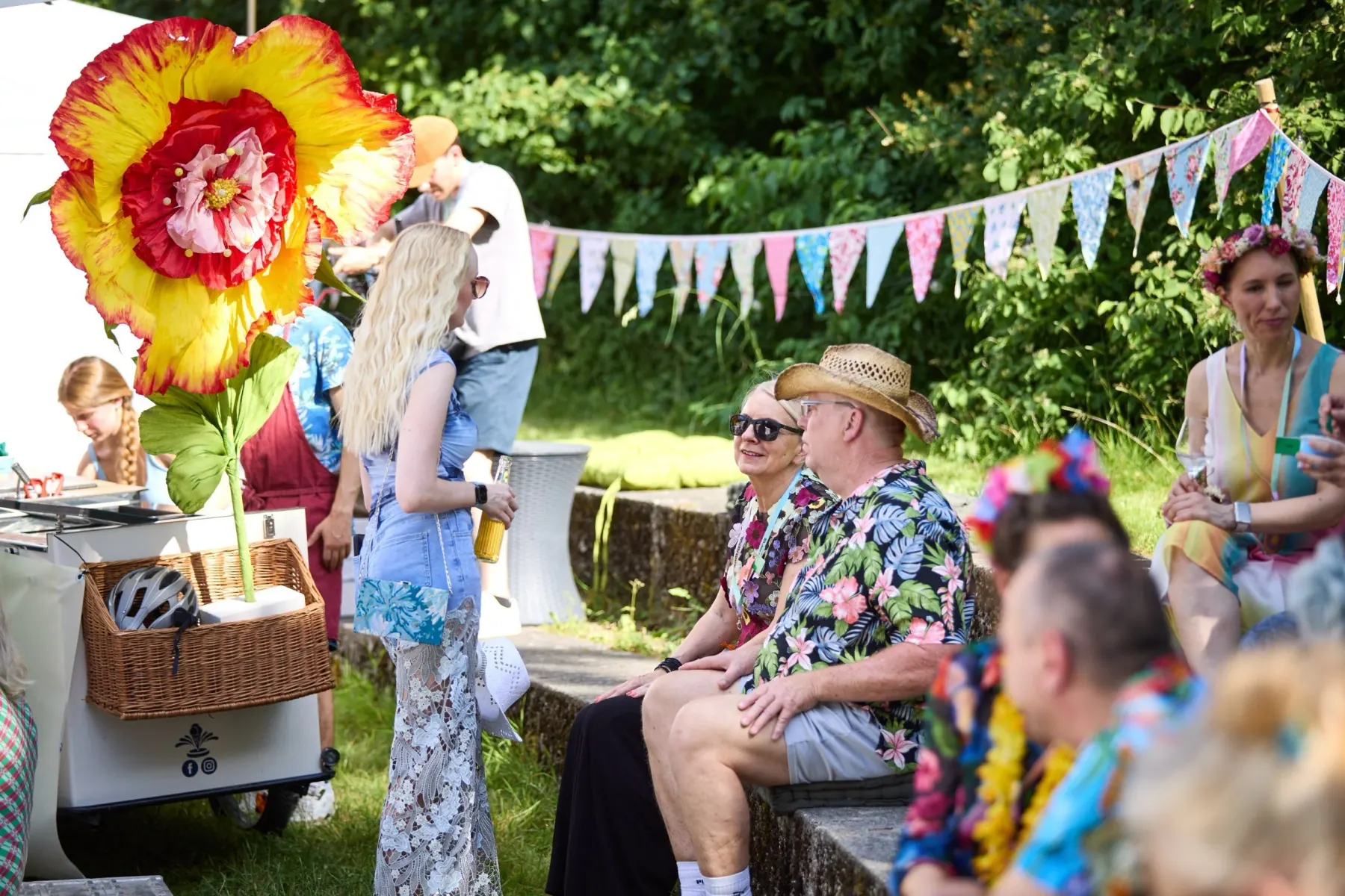 Guests in summer clothes sitting in the garden under bunting, an oversized paper flower standing to the left