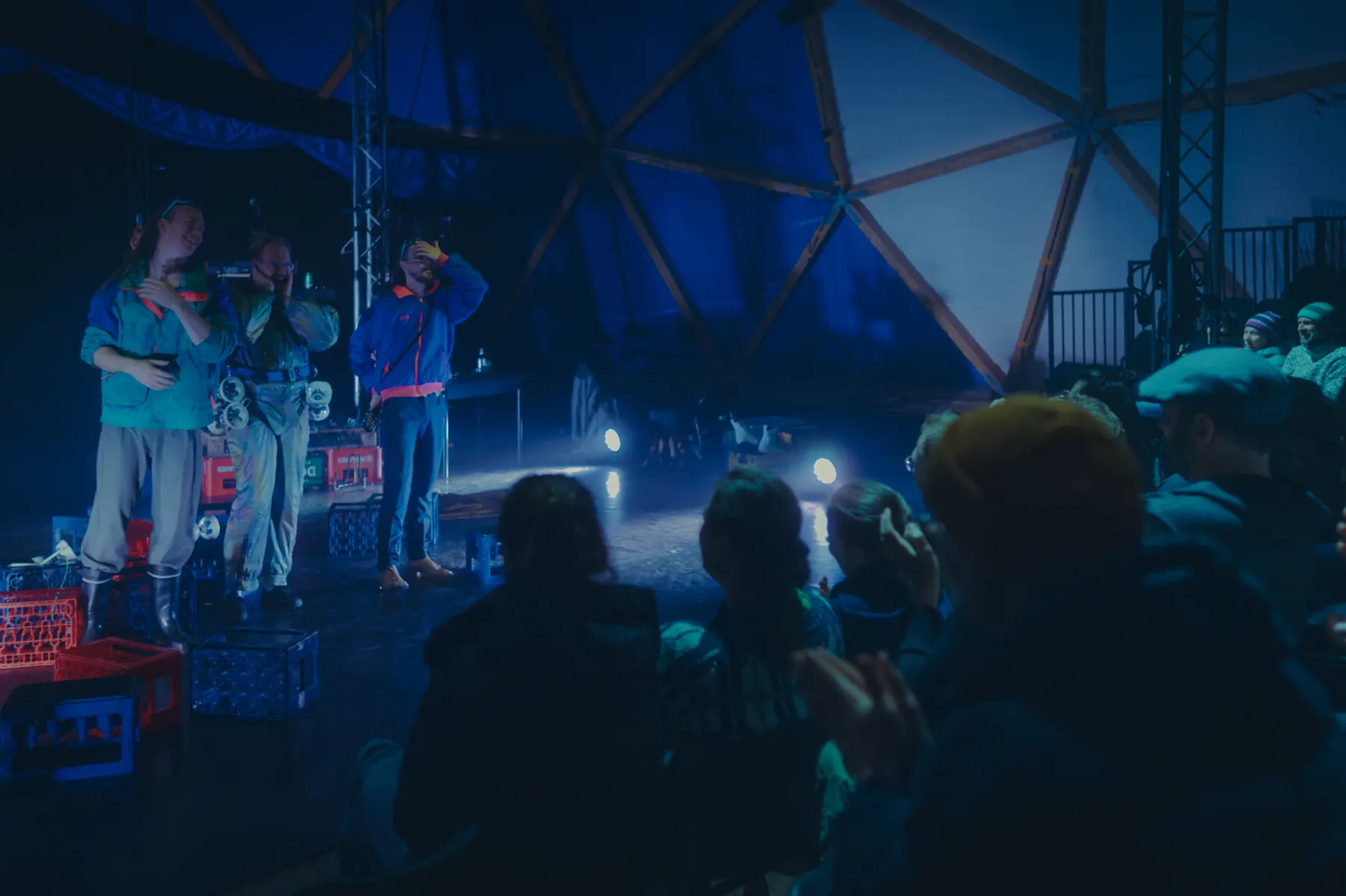 Three people standing on a stage built from drinks crates speaking into microphones, the audience seated in blue light