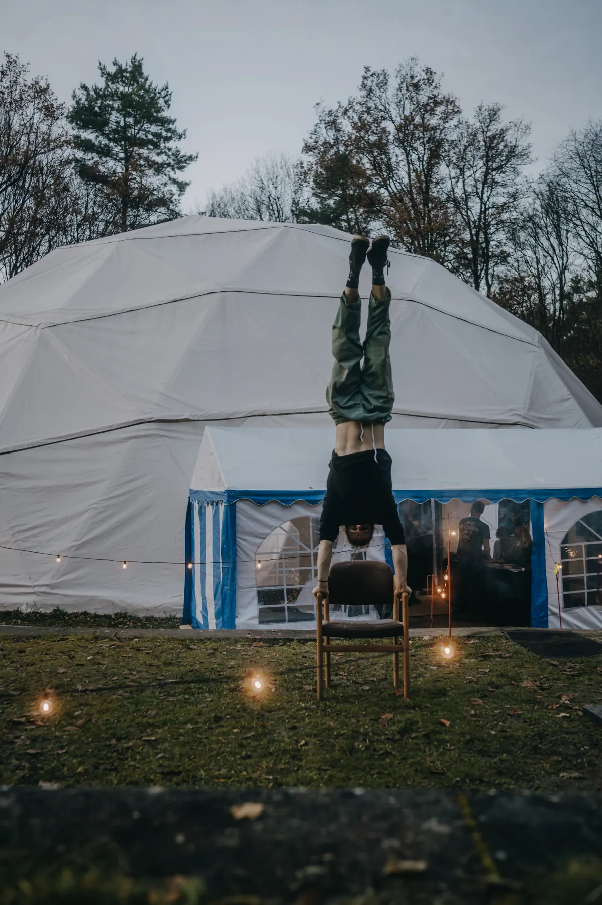 Artist in a handstand on the back of a chair in front of the lit dome, lanterns standing in the grass