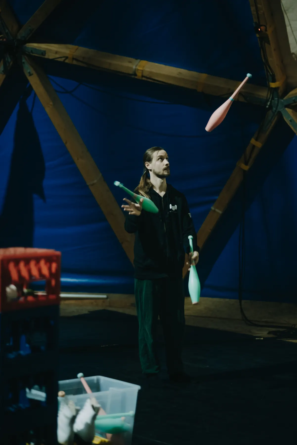 Juggler throwing clubs in the air, the blue canopy and wooden framework of the dome behind him