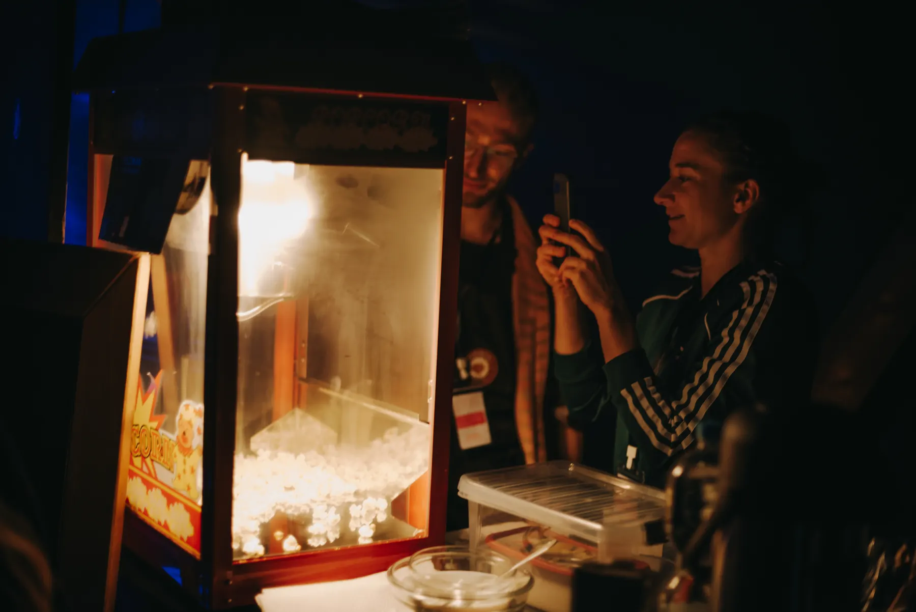 Two people at a lit popcorn machine, one of them filming with a phone