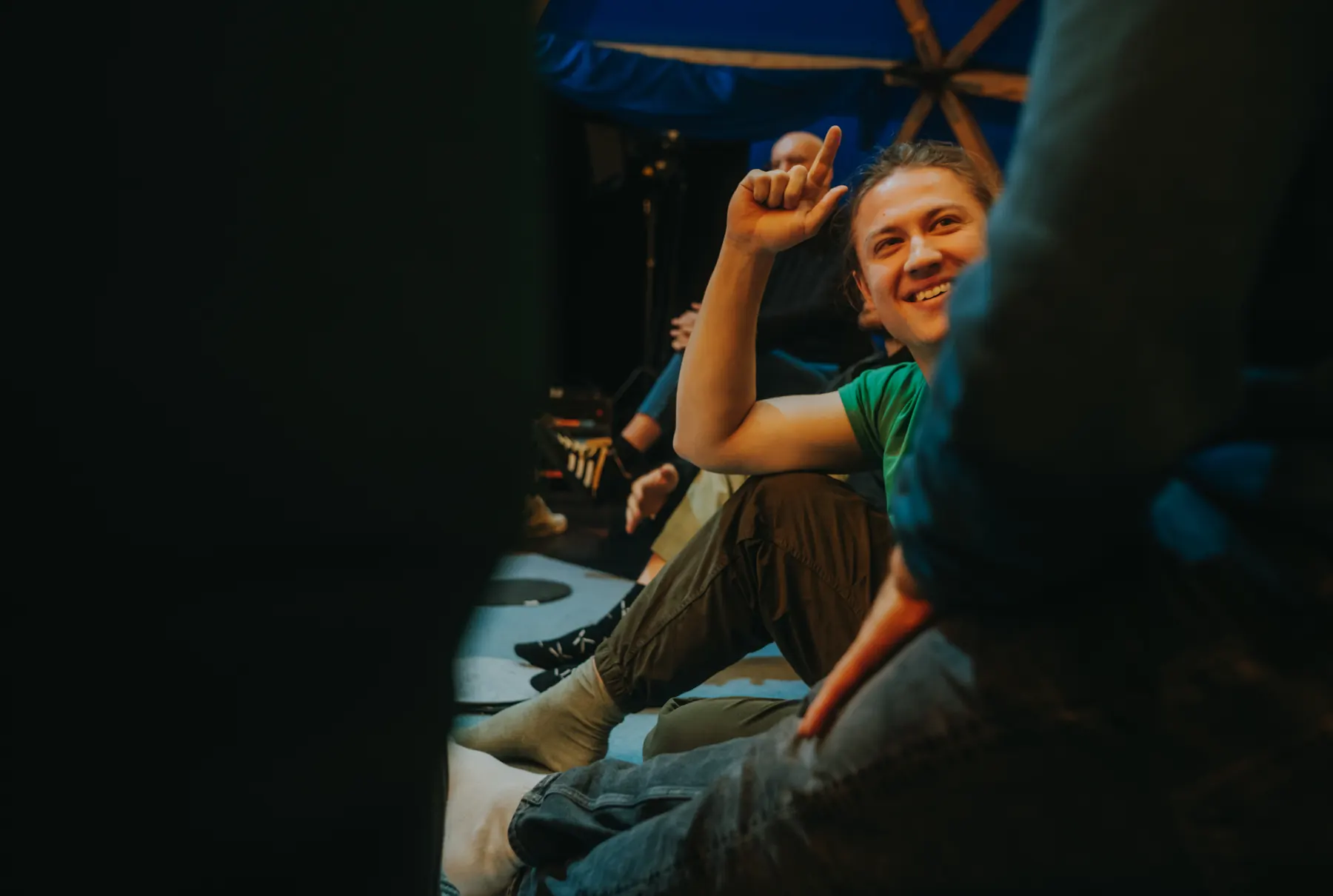 A man sitting on the floor among other guests, laughing and raising a finger