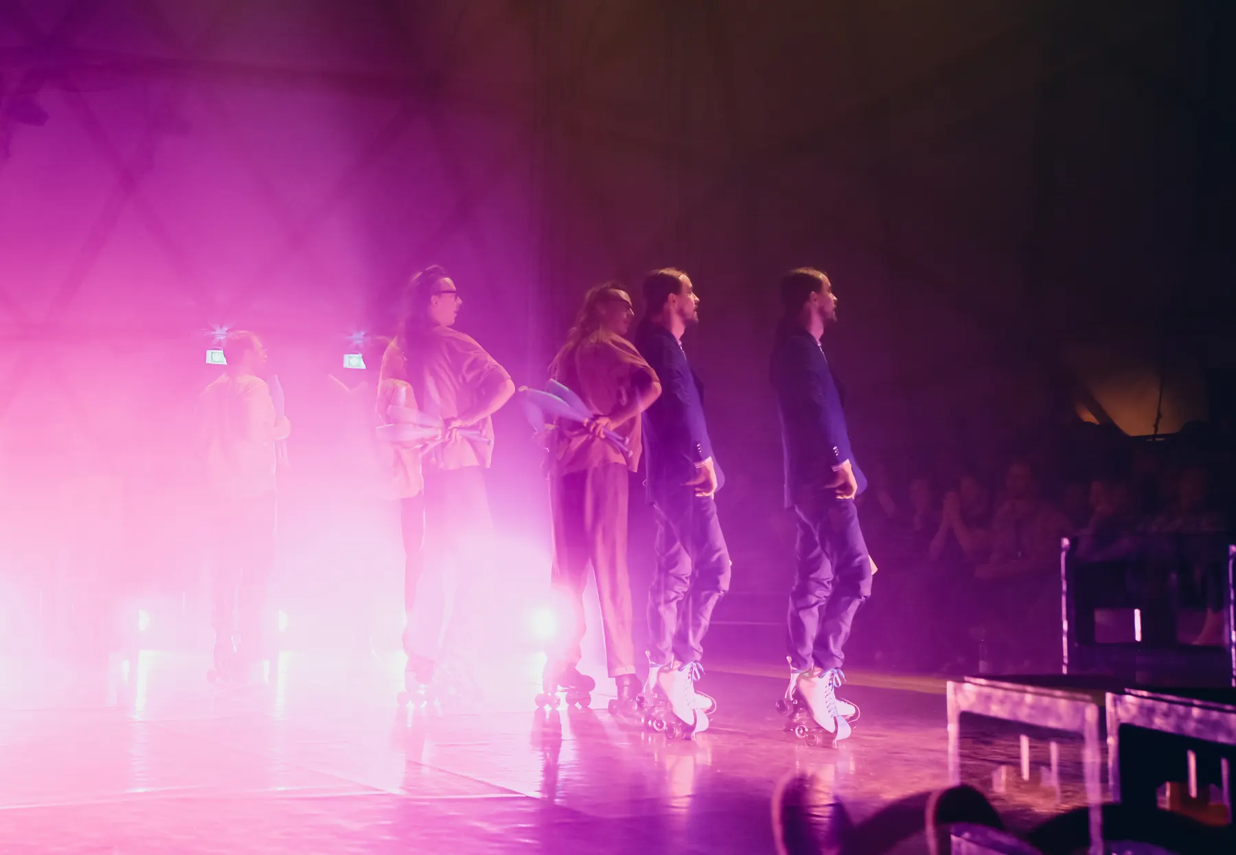 Five artists on roller skates standing side by side in pink haze, the audience in front