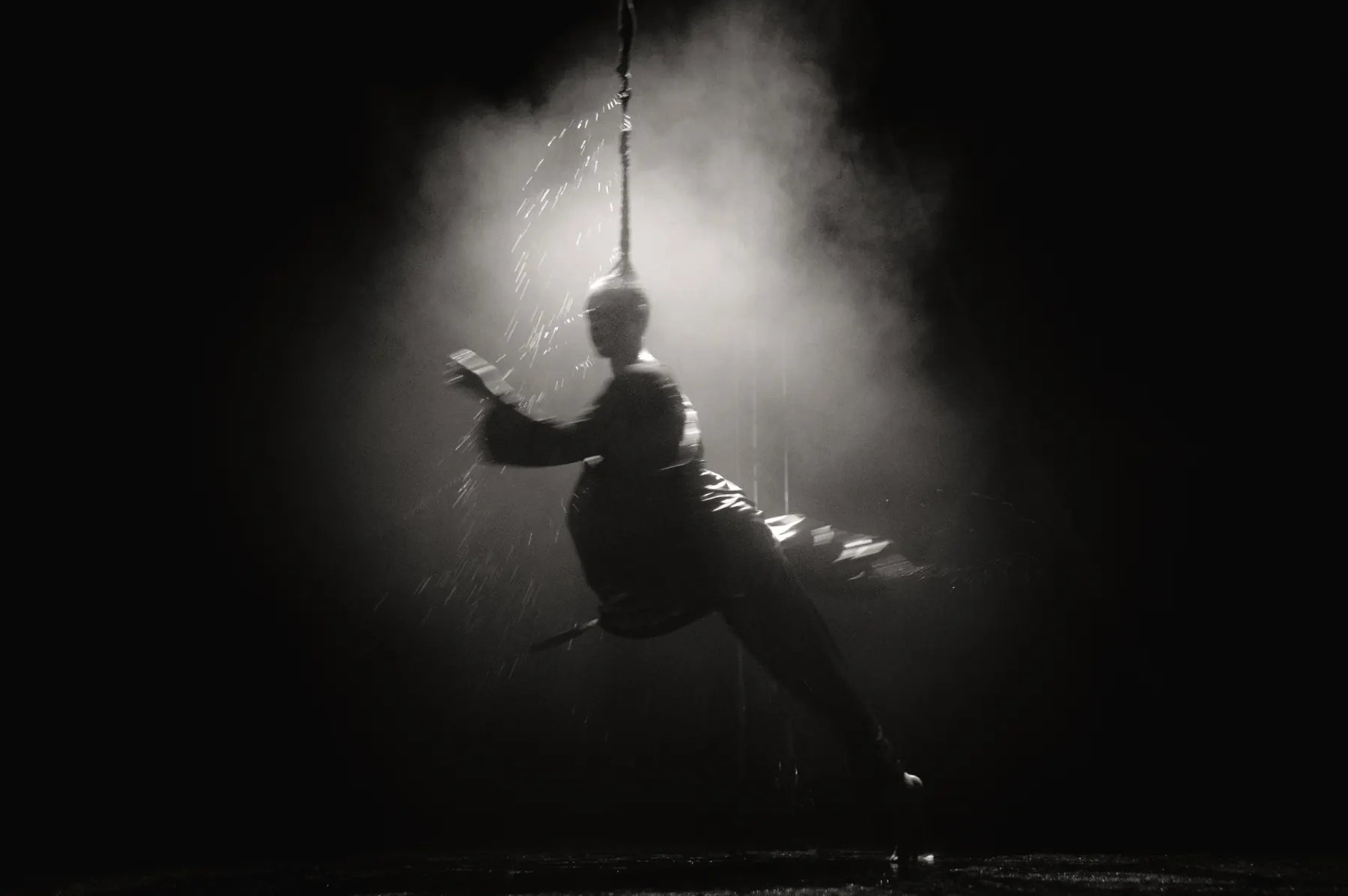 Artist hanging from a vertical rope, flinging drops of water through the lit haze