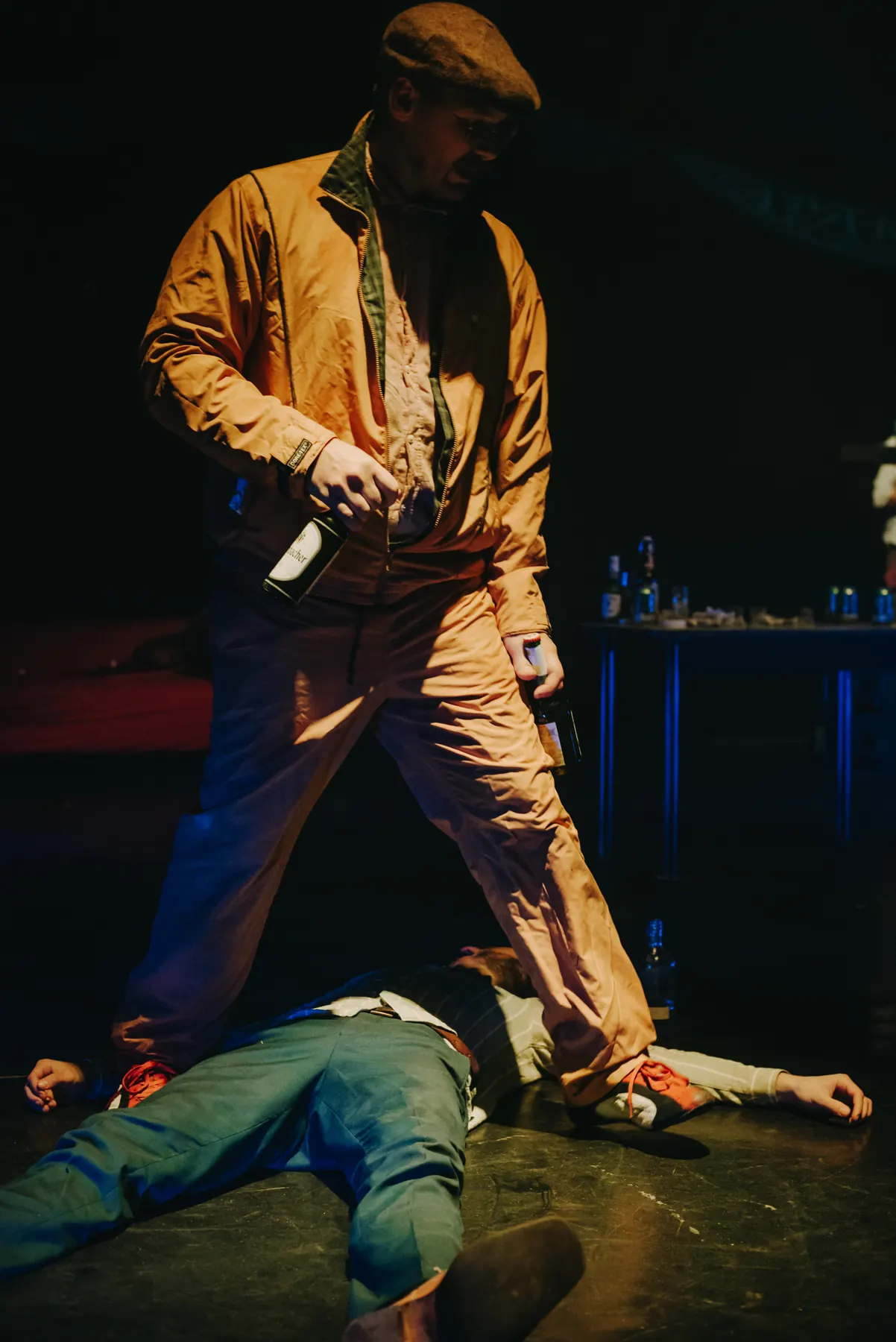 A performer in an ochre jacket and flat cap standing astride a person lying on the floor