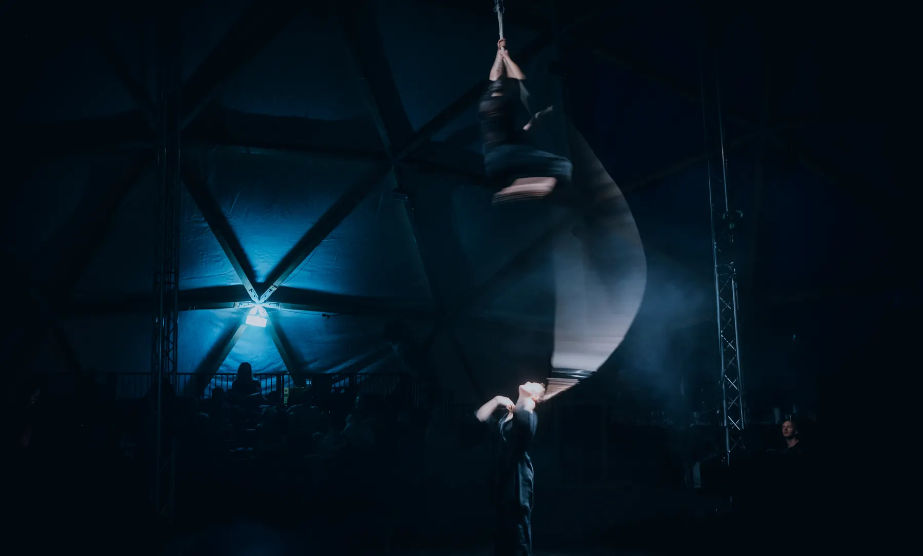 Artist high up on an aerial silk while a second person holds the fabric below, blue light shining through the dome frame behind