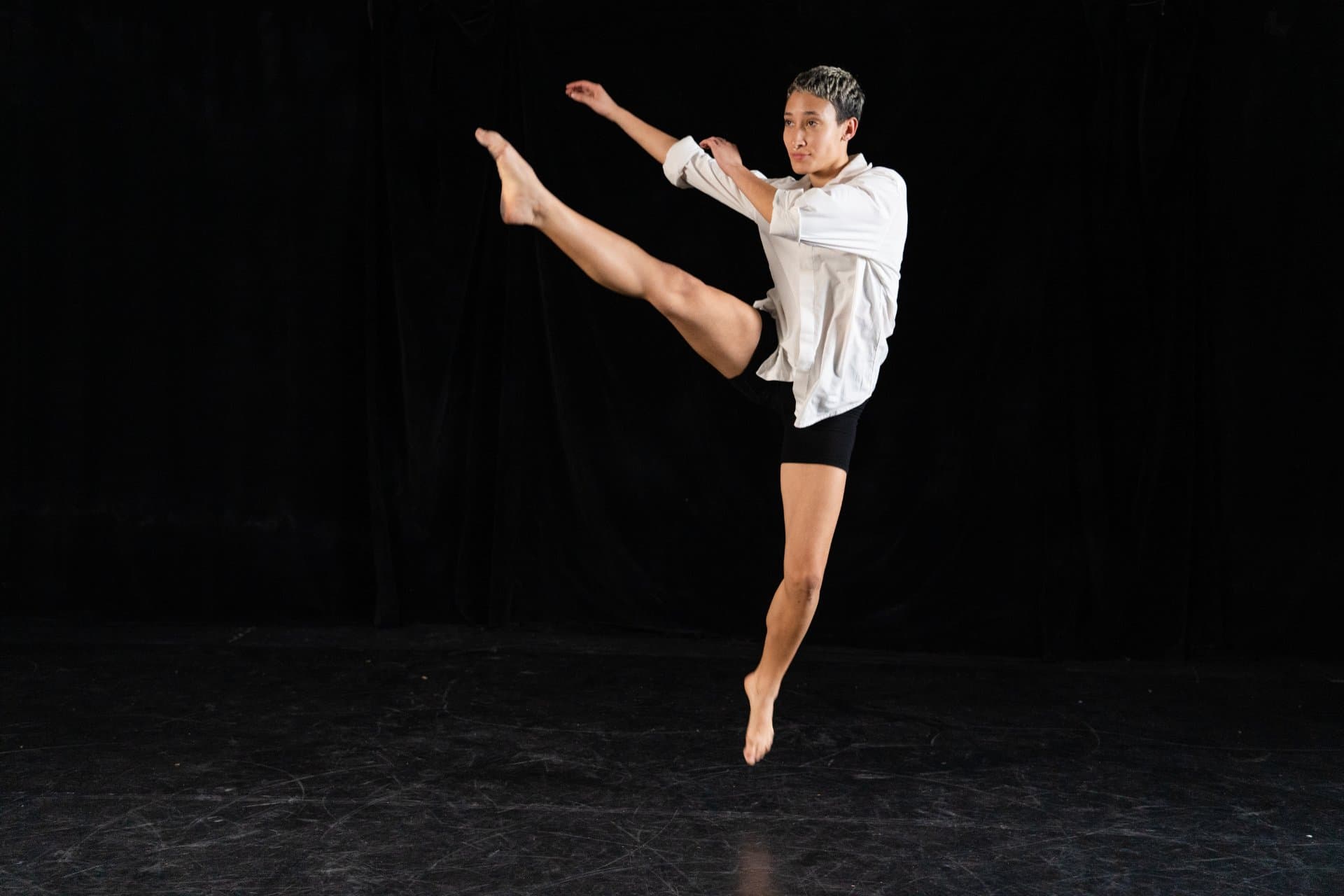 Dancer in a white shirt mid leap with an extended leg on a black stage