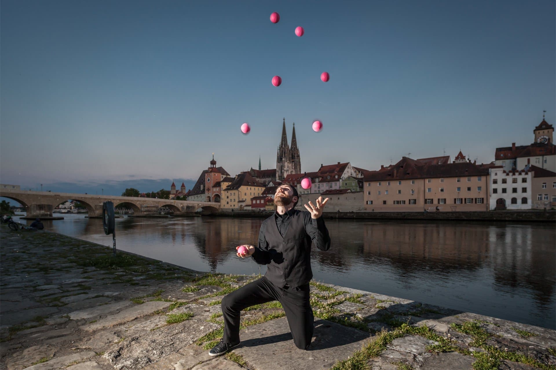 Juggler with five balls in the air on a riverbank, an old town with church spires behind